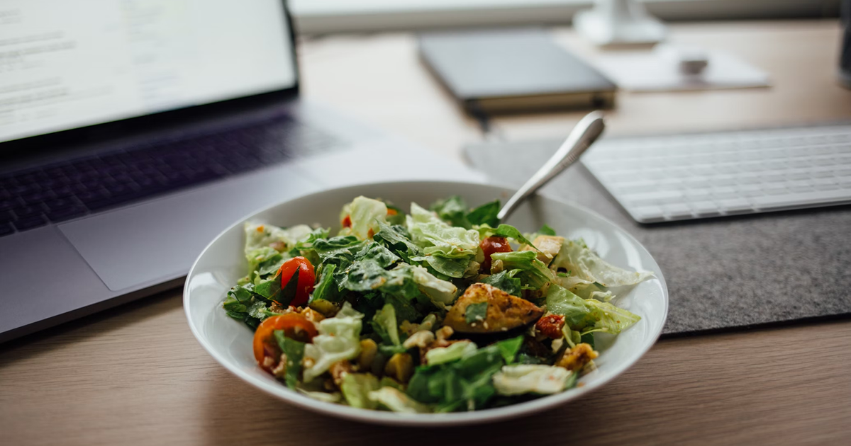 Salada fresca e colorida em um prato branco, localizada centralmente em uma mesa de escritório moderna, com um notebook aberto ao fundo, simbolizando um almoço equilibrado durante a jornada de trabalho.