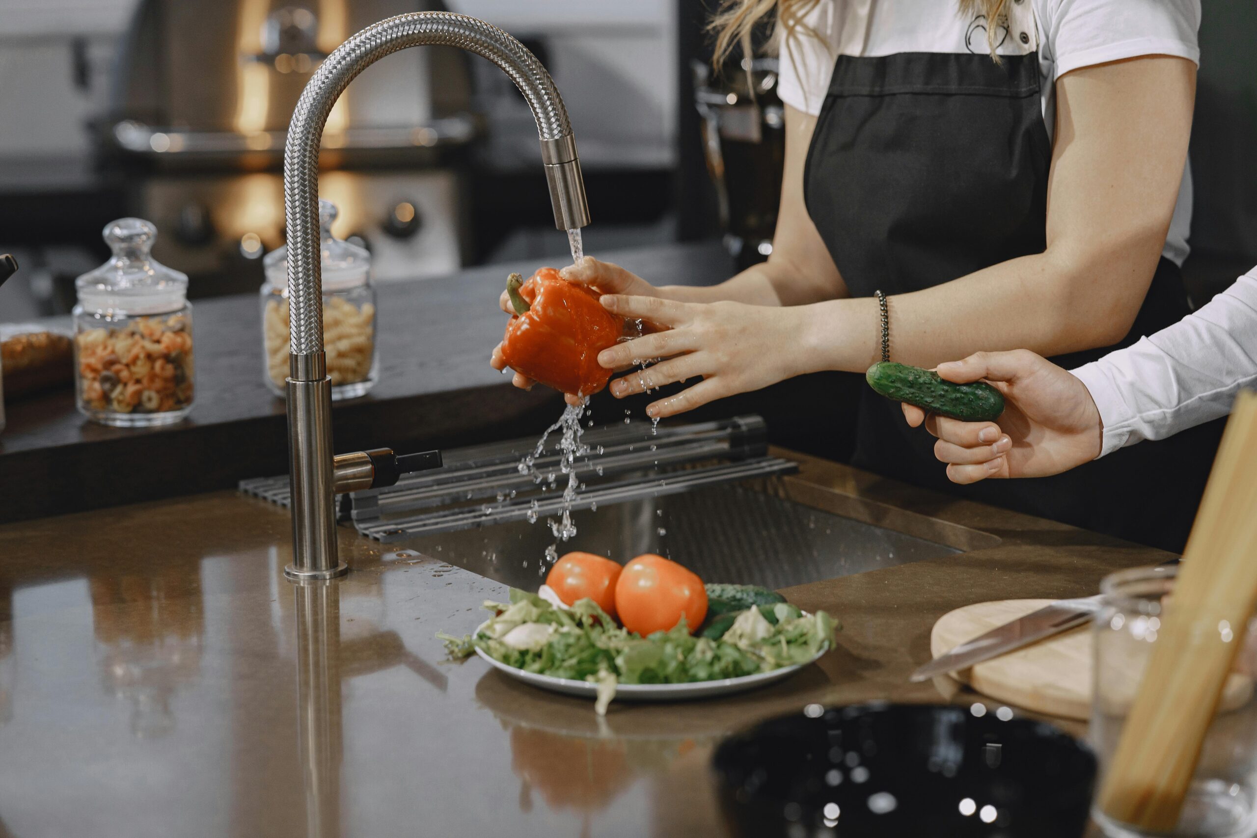 Duas pessoas lavando pimentão e pepino sob a água corrente em uma cozinha moderna, com alface e tomates frescos em um prato.